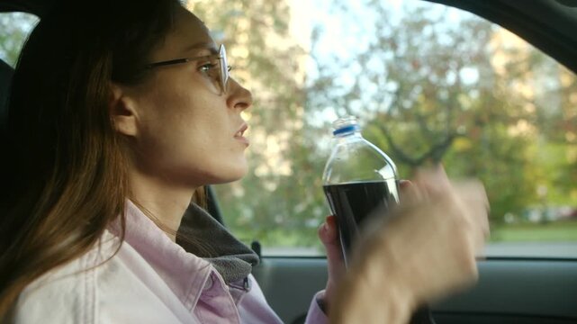 Young woman with glasses sitting in her car opening a plastic bottle and drinking a dark, sugary, carbonated beverage to quench her thirst while taking a break from driving on a sunny day