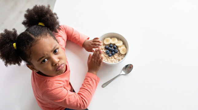 Pouting girl pushing away oatmeal bowl, picky eater at breakfast