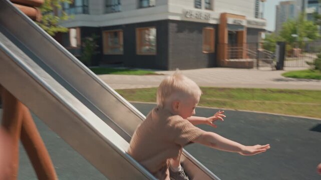 Young boy slides down metal slide at outdoor playground wearing casual clothing. woman crouches nearby to assist him as boy reaches bottom and then lifts and hugs him affectionately in front
