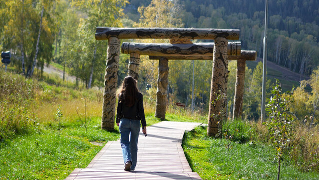 Rear view of a female tourist strolling on a mountain trail through traditional wooden gates in Altai in autumn