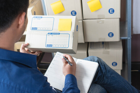 Man holding Cardboard Parcel Box and Writing on Notebook in Warehouse, Young Male Entrepreneur Checking Logistics Stock Order