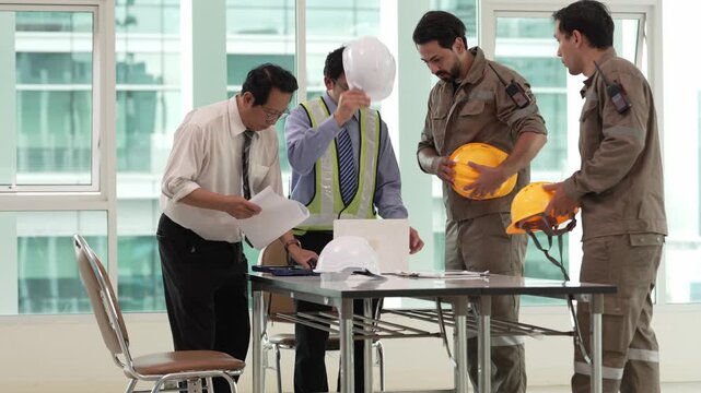 Engineering team reviewing construction plans around office table while wearing safety helmets