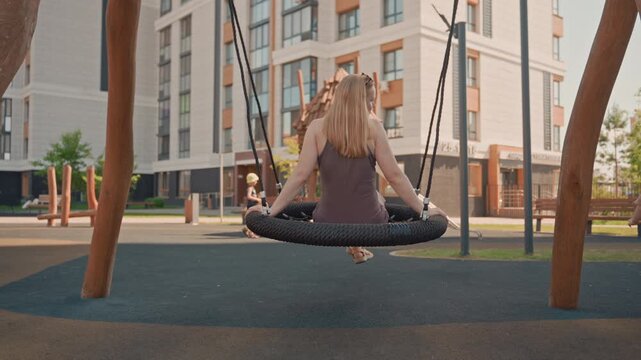 Woman enjoys swinging on tire swing at outdoor playground with her young son nearby. scene features modern apartment buildings in background wooden play structures including slides