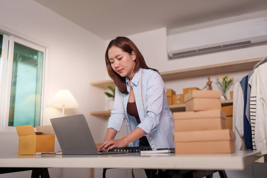 Asian woman entrepreneur using laptop to manage online store orders near cardboard boxes