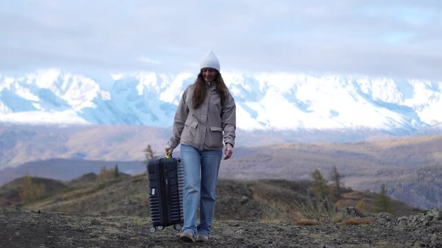 Smiling happy woman tourist pulling a suitcase and walking forward against a snow covered mountain range with a slow camera push in.