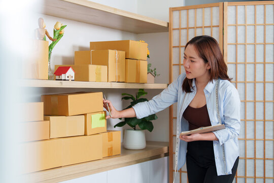Asian businesswoman holding clipboard while checking cardboard stock boxes on shelves