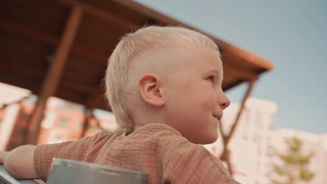 Young boy with short blonde hair sits on swing facing away from camera towards tall apartment buildings. woman stands nearby smiling and clapping in support as boy enjoys outdoor playtime