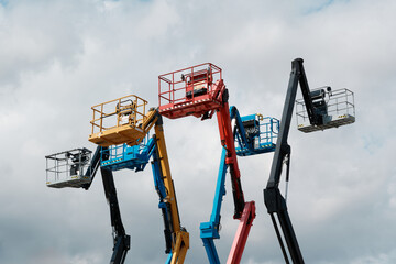 Row of colorful telescopic boom lifts and cherry pickers with raised working baskets against cloudy sky. © Ekaterina