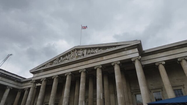 A Union Jack flies above a sculpted pediment and stone columns beneath a cloudy sky