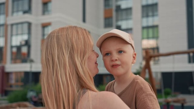 Woman and young boy interact closely in outdoor urban setting with background featuring modern buildings and playground. scene is calm showing their connection against soft daylight