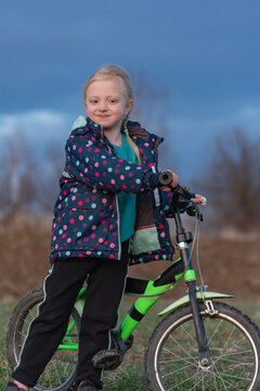 Blonde girl with braid smiling at camera, standing proudly beside a green and black bicycle on grass during an outdoor cycling moment of childhood fun and growth