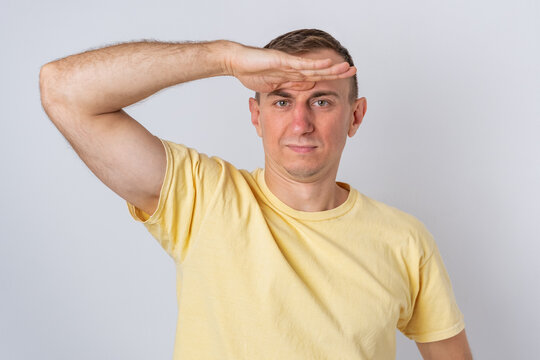 Young man wearing a yellow t shirt looking intently into the distance, shielding his eyes from bright light with his hand, searching for something or observing the horizon