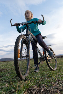 Blond boy standing with a mountain bike in a grassy field, holding the handlebars, looking forward with focus under a bright sky, emphasizing youth and outdoor activity