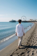 Fototapeta premium Mature woman walks alone on dark sand beach facing blue ocean. Wears white shirt, light pants for summer vacation. Seaside pier with ferris wheel stands in background under clear sky. Peaceful