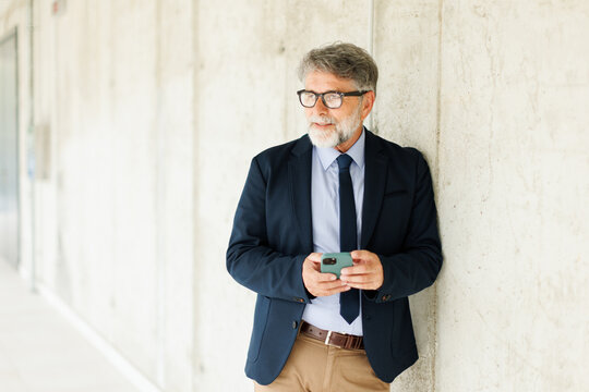 Confident businessman holds a smartphone against a concrete wall