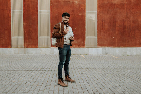 A smiling father stands outdoors wearing his baby boy in a carrier