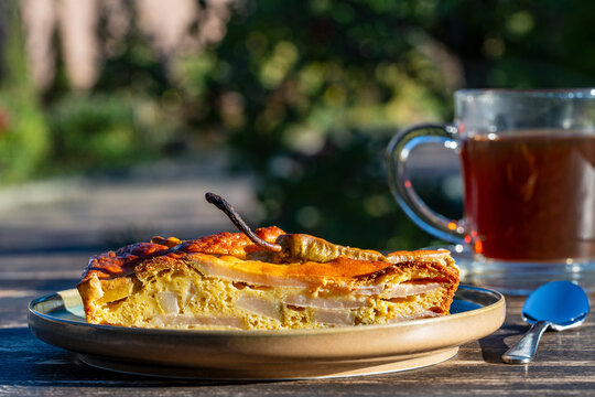 Slice of homemade pear pie with cup of tea. Closeup of delicious piece of pear cake served on plate with glass mug of hot tea on wooden table outdoors in sunny garden