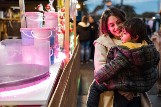 Mother holding toddler boy at illuminated cotton candy stall at night