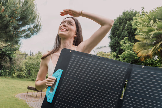 Smiling woman holds a portable solar panel in her sunny home garden