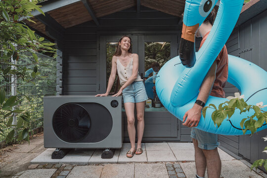 Woman and man with flamingo float by heat pump outside poolhouse