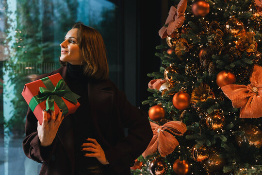 Smiling woman holds wrapped gift near decorated Christmas tree