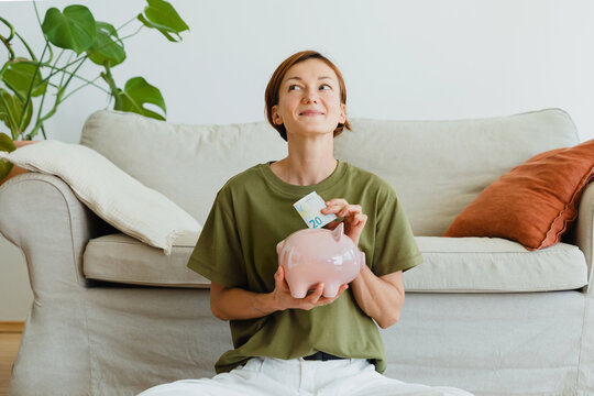 A smiling woman puts money into a piggy bank at home
