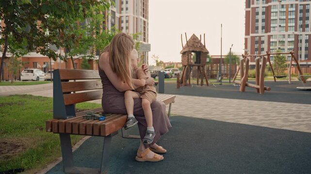 Woman sits on bench in urban park holding and comforting young child. scene includes toy vehicles placed beside them with playground equipment and residential buildings visible in background