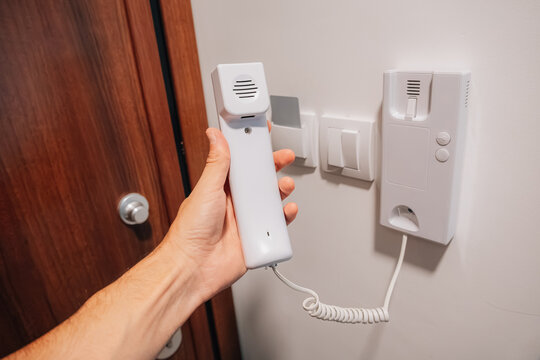 Home security and apartment access concept: hand holding intercom handset lifted from wall-mounted unit near front door.