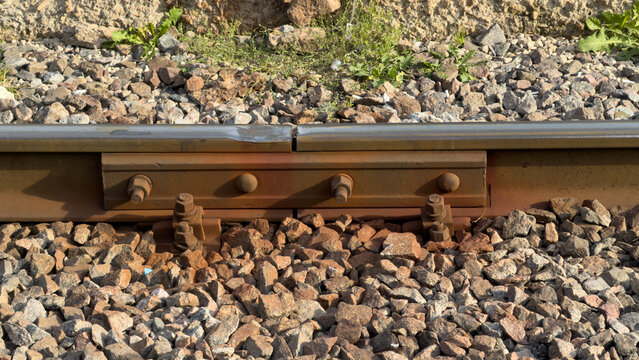 Rail track with bolts and gravel bed. Sunlight highlights metal and stones. Close-up view shows rail joint details. Rust and wear visible on track.