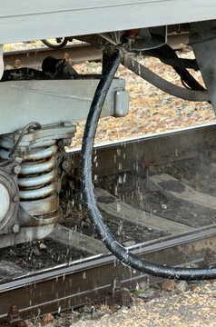 Water drips from a train's undercarriage. Metal pipes run beneath the rail vehicle. Dirt and gravel cover the train tracks.