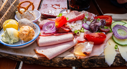 Close view of traditional Austrian Bavarian Brettljause alpine snack board served on a wooden platter, with cured meats, sausages, cheeses, tomato, cucumber, red onion and mini pretzel © Peter Stein