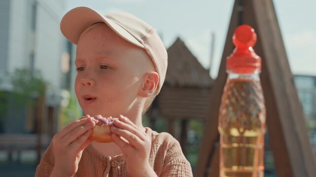 Young boy enjoys eating donut while wearing beige cap and brown textured shirt. large plastic bottle with red lid is visible in background along with outdoor playground equipment