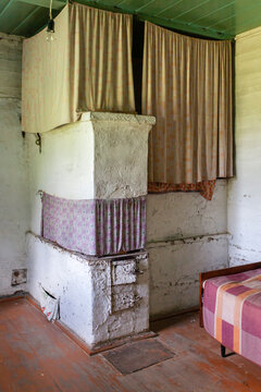 Interior of old rustic house featuring a traditional white plastered wood-burning stove decorated with vintage fabric curtains. Atmosphere of traditional rural lifestyle and authentic interior design.