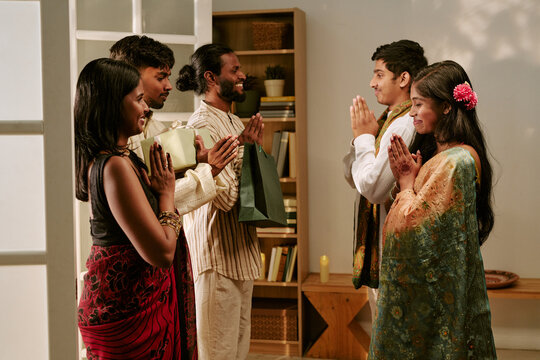 Young adult Indian friends greeting each other with namaste gesture, holding gift boxes indoors. Visual representing cultural holiday traditions for festive marketing campaigns