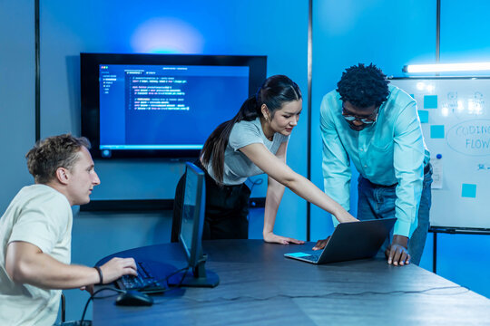 Diverse programming team collaboration in modern blue-lit tech lab. Woman and man analyze code on laptop, while another developer observes. Focus on innovation, teamwork, and software development flow