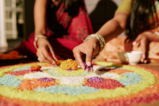 Cropped hands of Indian young adult women creating colorful floral rangoli on floor during festival celebration. Visual asset representing traditional culture for holiday marketing