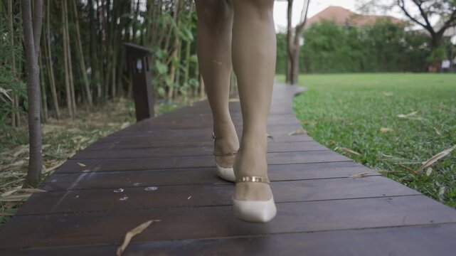 A woman in elegant high heels walks gracefully along a wooden pathway in a lush garden. Her crossed footsteps create a stylish and confident stride.