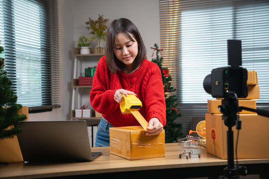 Asian woman packing a cardboard shipping box with adhesive tape in a room with Christmas decorations after livestream