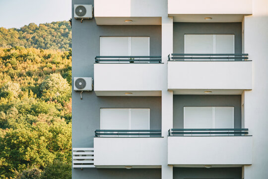 Residential property listing or real estate: modern apartment block facade with balconies and AC units, wooded hillside backdrop.