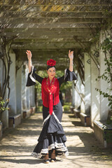 An Andalusian woman wearing a black flamenco dress and a red shawl, with flowers in her hair, dancing flamenco under a colonnade covered in climbing plants in a park. Concept of dance, art. © Manuel