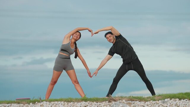 Two people stretch together, forming a heart shape. They stand on a pebbled beach, sky behind them. Their smiles show happiness and connection