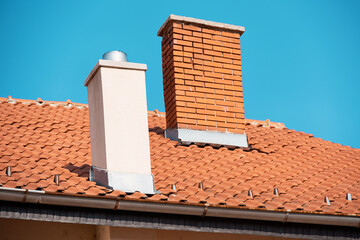 Obraz premium Two chimneys on a clay tile roof with metal flashing and rain gutter against clear blue sky.