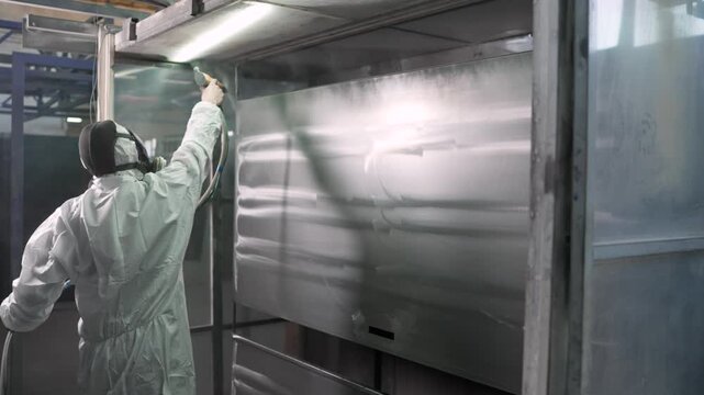 Industrial painter in full hazmat gear applies finish to metal component inside spray booth. Worker holds electrostatic gun, covering surface with fine powder. Professional manufacturing work