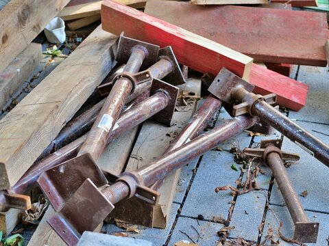 Rusty adjustable steel trench props, screw jacks, and wooden beams stored outdoors on paved ground at construction equipment site.