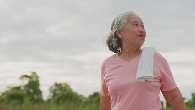 Elderly woman stretches in park, smiling, arms outstretched. She wears pink shirt, towel around neck. Cloudy sky, green trees in background