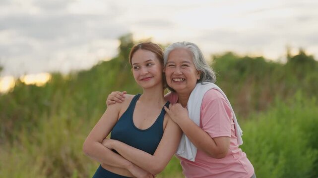 Golden light warms two smiling women embracing in a field. Younger woman wears athletic gear, arms crossed, looking happy. Older woman laughs