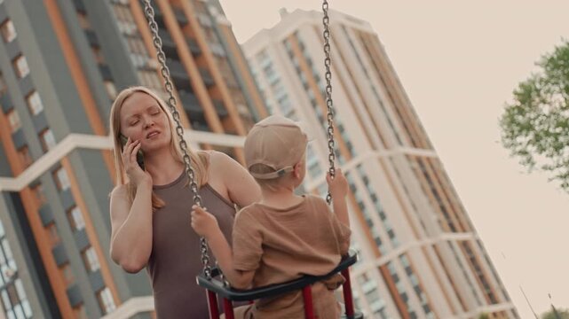Woman talks on her phone while young boy swings at outdoor playground with tall buildings in background. scene captures everyday life moments focusing on interaction between mother and child