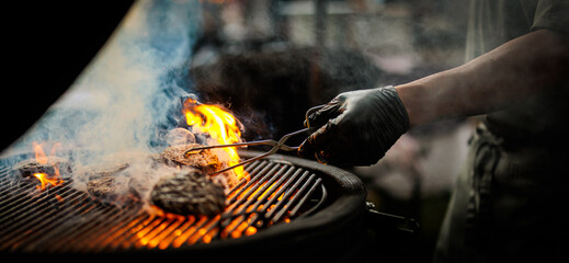 Close-up of a chef grilling beef burgers over open flames on a barbecue, with dramatic fire, smoke and glowing heat creating an atmospheric outdoor cooking scene. © Sjors