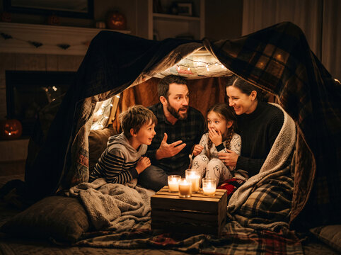 Whispering parents and kids sharing scary tales in blanket den with lanterns
