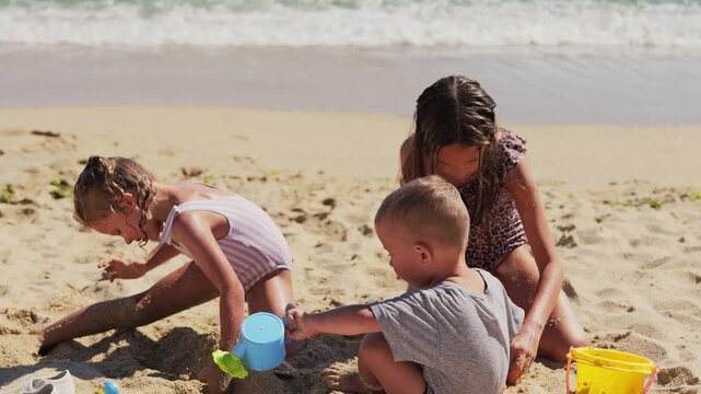 Three young children playing together in golden sand with colorful buckets and beach toys, enjoying summer family vacation outdoors near the sea.